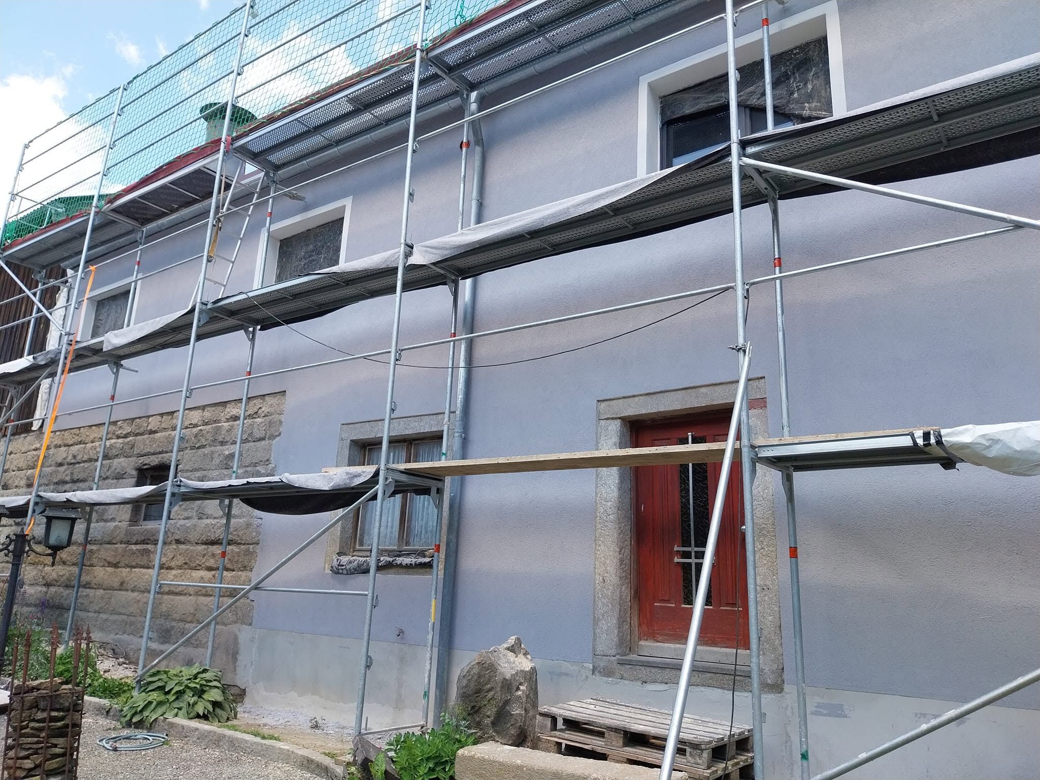Eingerüstete Hausfassade mit heller Putzfläche, Natursteinsockel und roter Haustür
